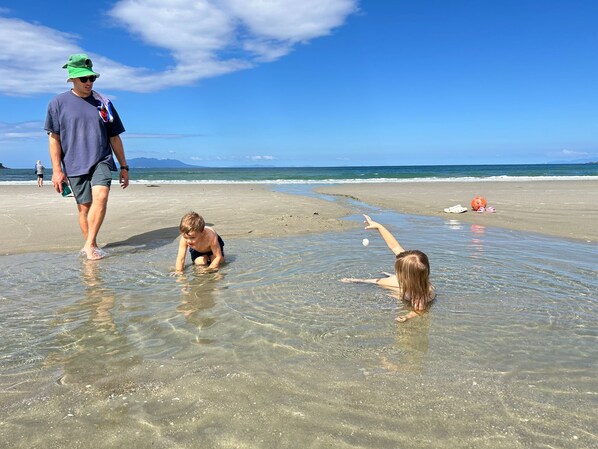 Een strandscène met helder ondiep water, een persoon die loopt en twee kinderen die aan het spelen zijn.