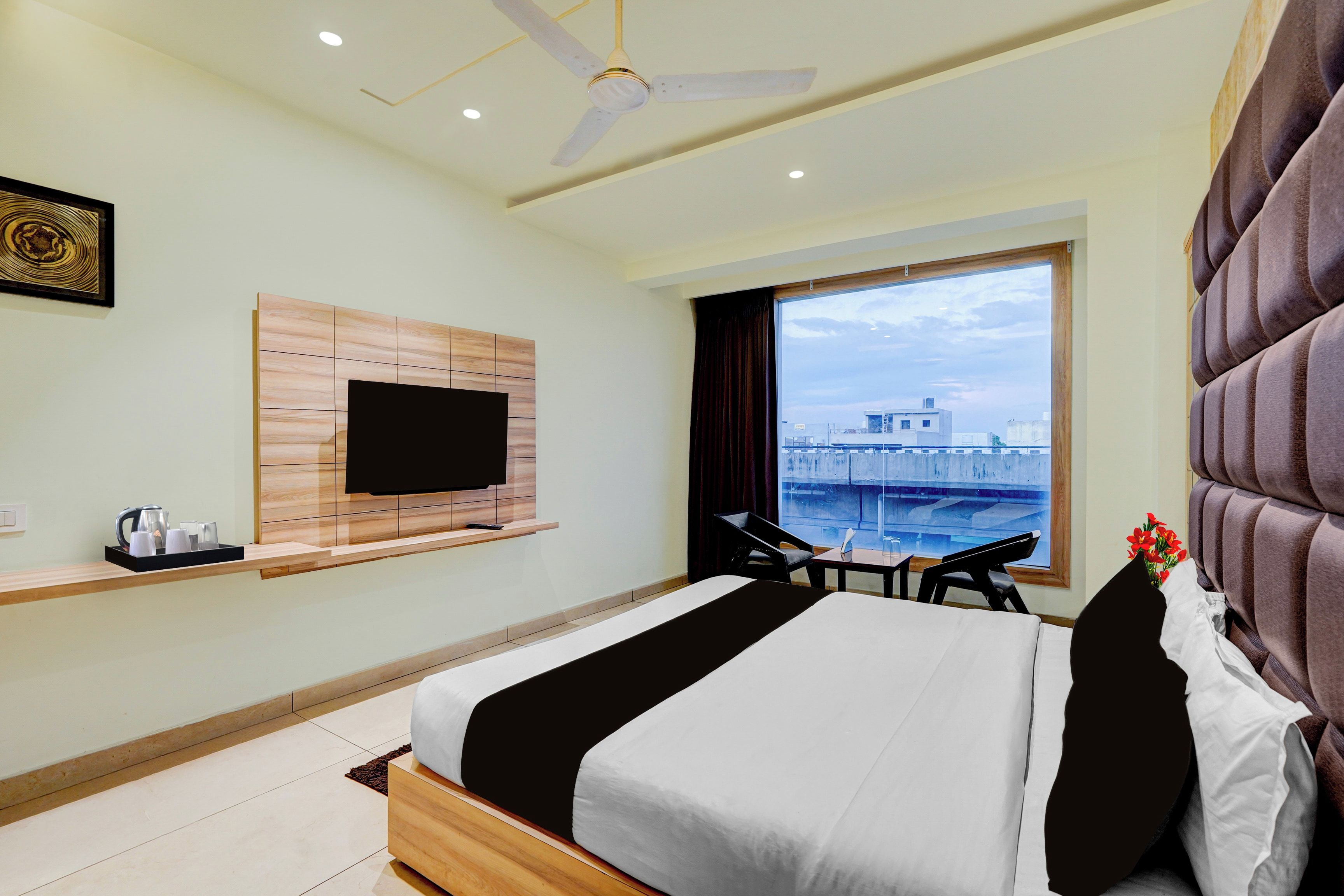 Ein Hotelzimmer mit einem Bett, einem flachbildfernseher an einer Holzvertäfelung, einem Fenster mit Blick auf ein Gebäude und einem Deckenventilator.