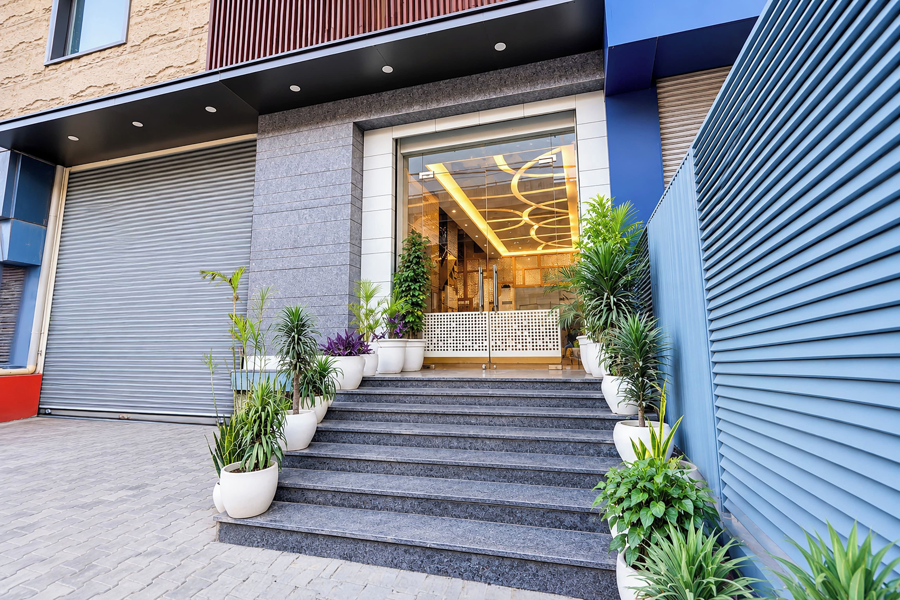 A building entrance with a set of steps leading up to a glass door, flanked by potted plants and a closed garage door.