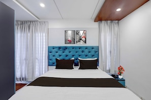 A modern bedroom with a tufted headboard, a large bed, two black pillows, white bedding, and a nightstand with a phone and a vase of flowers. - Townhouse Karur Bus Terminal Formerly (Karur)