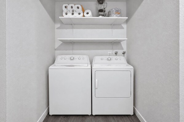 Ein Waschraum mit zwei weißen Waschmaschinen und einem darüberliegenden Regal mit Toilettenpapierrollen und einem Mixer.