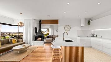 A modern living room with a fireplace, a kitchen with white cabinets, and a view of the outdoors.