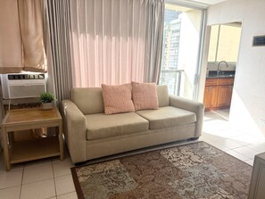 A living room with a sofa, a small side table, a potted plant, and a large window with curtains. - Prime Waikiki 1BR | Free Parking, Pool & W/D (Honolulu)