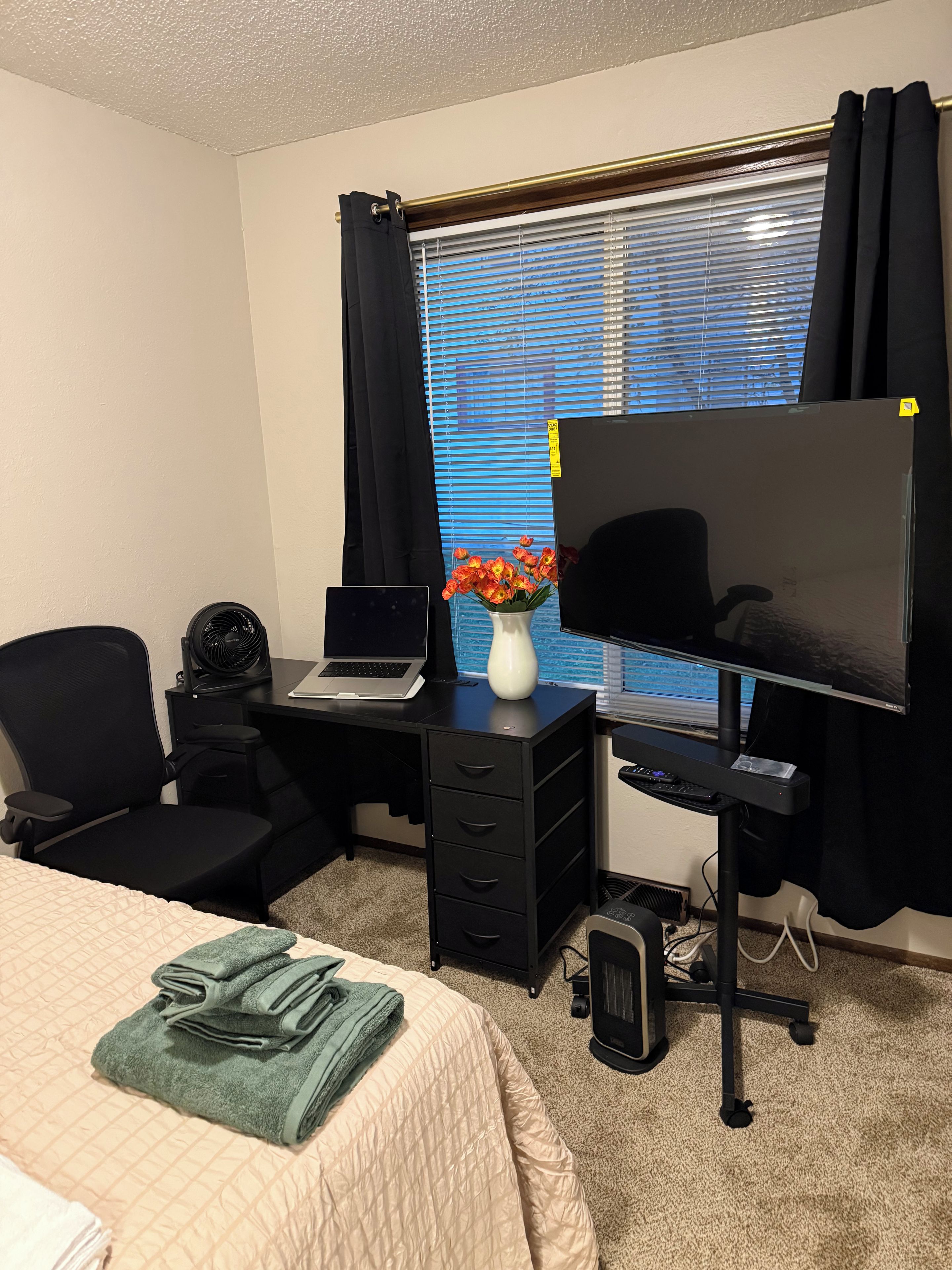 A hotel room with a bed, a desk with a laptop, a TV, a chair, a fan, and a window with curtains.