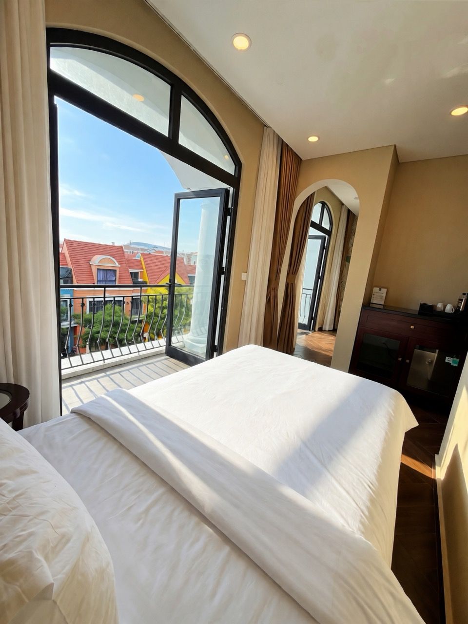 A hotel room with a large bed, a desk, and a view of buildings through a large window.