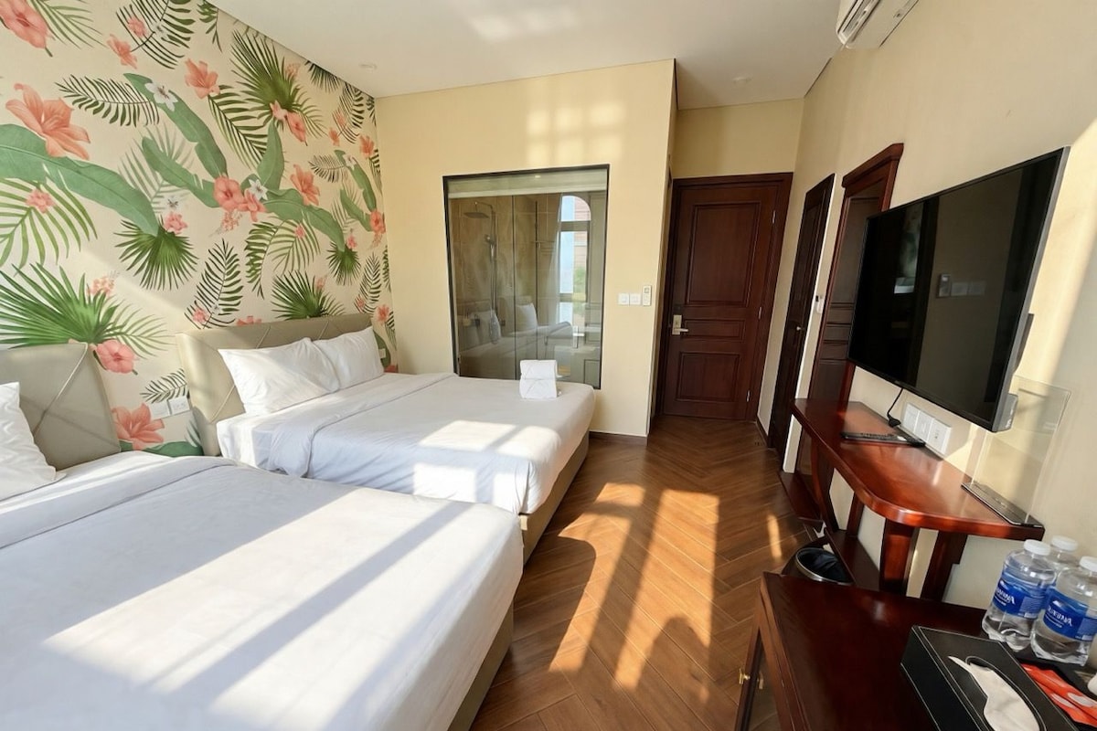 Habitación de hotel con dos camas, zona de ducha, televisor y escritorio con silla.