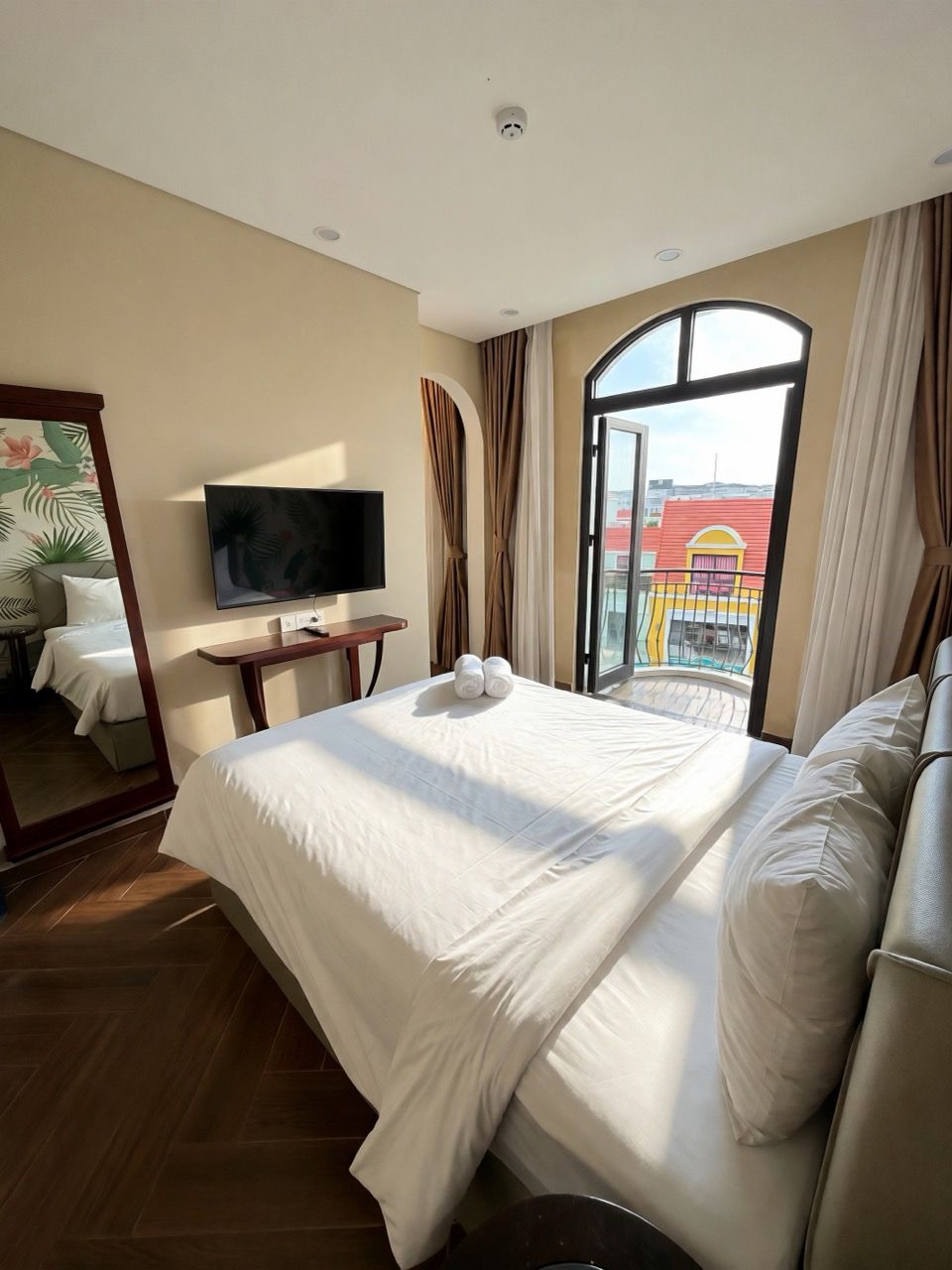 A hotel room with a bed, a TV, a window with curtains, and a view of buildings outside.