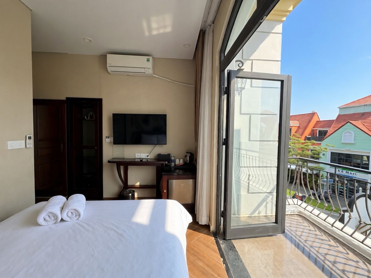 A hotel room with a bed, a desk, a TV, and a balcony with a view of buildings.