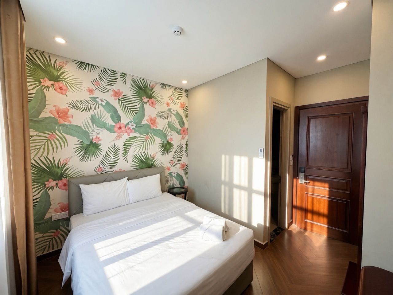 A bedroom with a bed, a nightstand, a wall with a tropical leaf and flower pattern, and a wooden door.