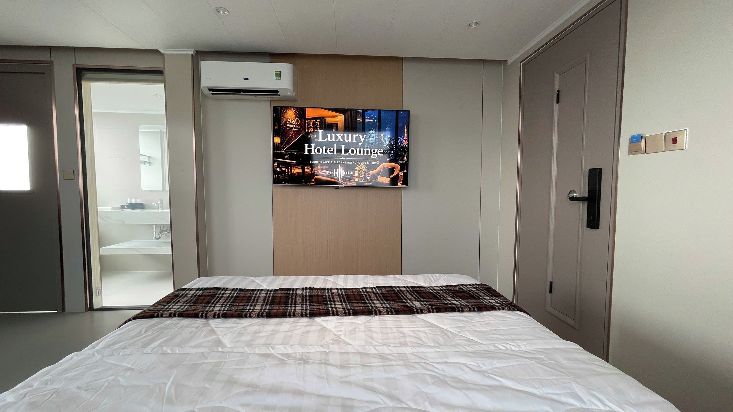 Una camera d'albergo con un letto, un condizionatore d'aria a muro, un quadro che raffigura un lounge dell'hotel e una porta con un interruttore.