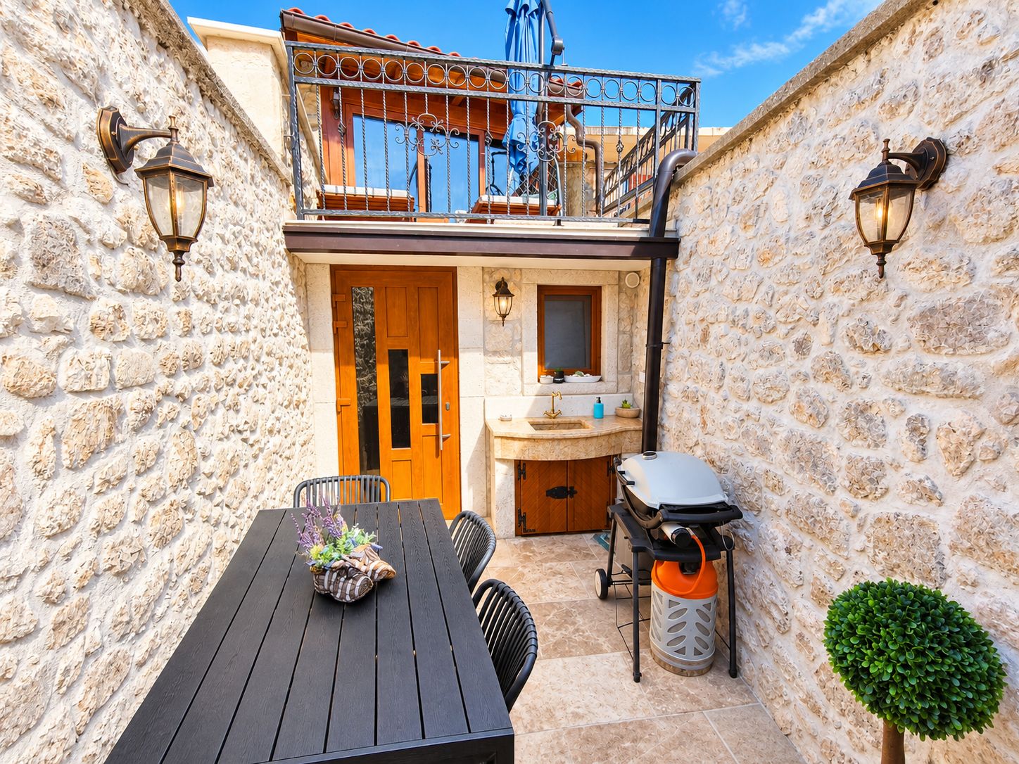 A stone-walled outdoor area with a wooden table, chairs, a barbecue grill, and a small potted plant.
