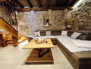A cozy room with a wooden staircase, a stone wall, a sectional sofa, and a wooden table with a candle and remote. - Villa Stone Retreat (Grižane-Belgrad)