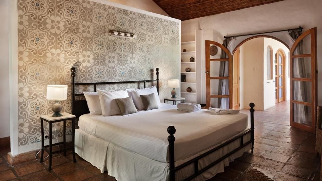A bedroom with a four-poster bed, a nightstand with a lamp, a wall with patterned wallpaper, and an arched doorway.