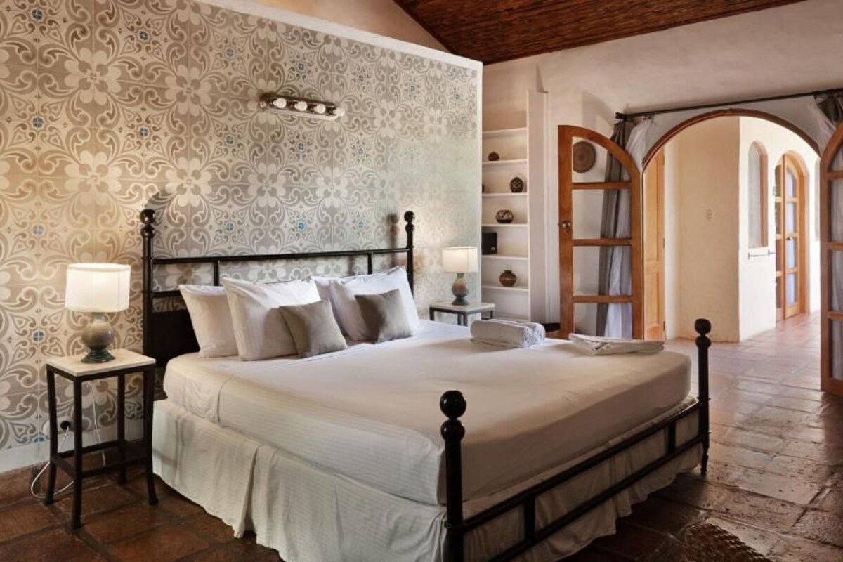 A bedroom with a four-poster bed, a nightstand with a lamp, a wall with patterned wallpaper, and an arched doorway.