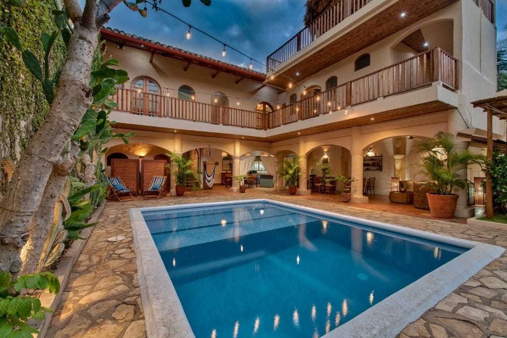 Zona de piscina con suelo de piedra, rodeada de plantas en macetas y mobiliario de exterior.