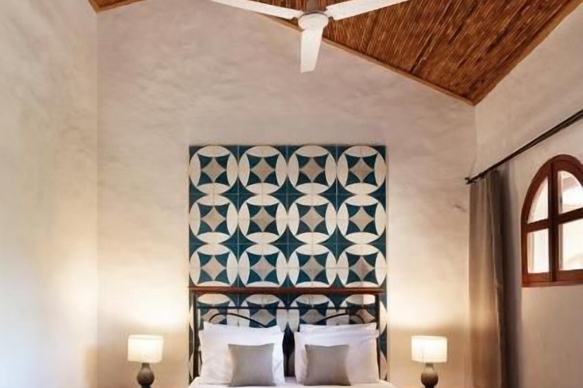 A bedroom with a large bed, a ceiling fan, patterned wall headboard, and two bedside lamps.