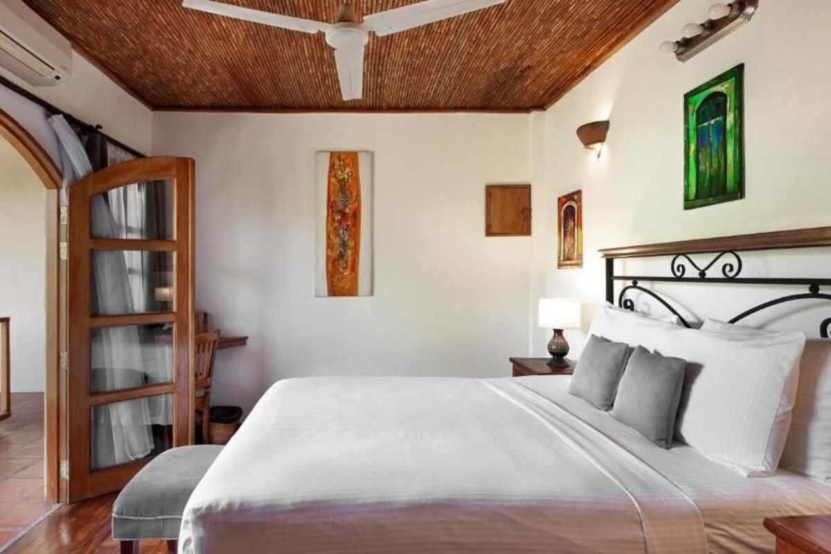 A bedroom with a bed, a wooden headboard, a nightstand, a wall-mounted lamp, and a painting.