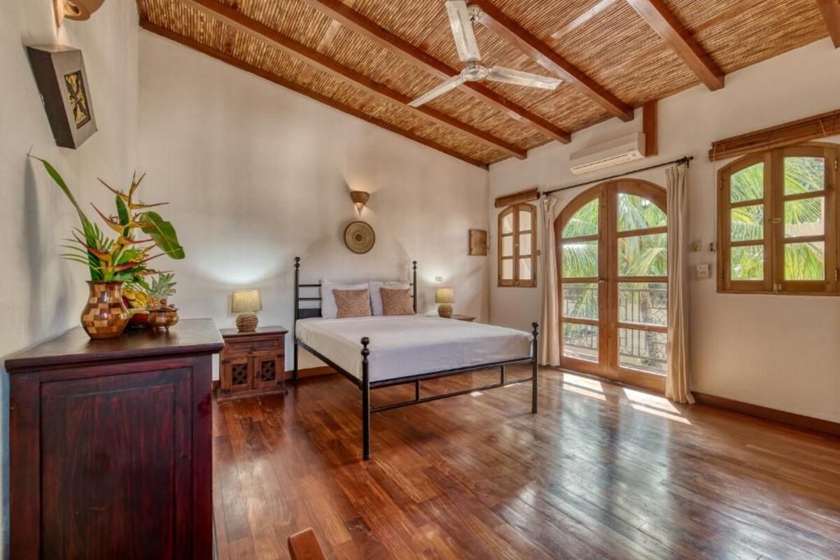 A bedroom with a bed, wooden flooring, a ceiling fan, and large windows.