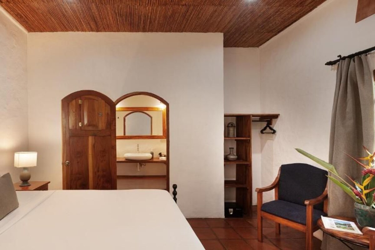A bedroom with a bed, a wooden wardrobe, a chair, a bedside table, a mirror, and a shelf with items.