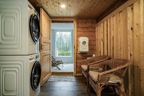 A laundry room with stacked washing machines, wicker chairs, a drying station, and a view through a door to an outdoor area. - Arctic Sauna & Aurora Retreat By Lapland Host (Rovaniemi)