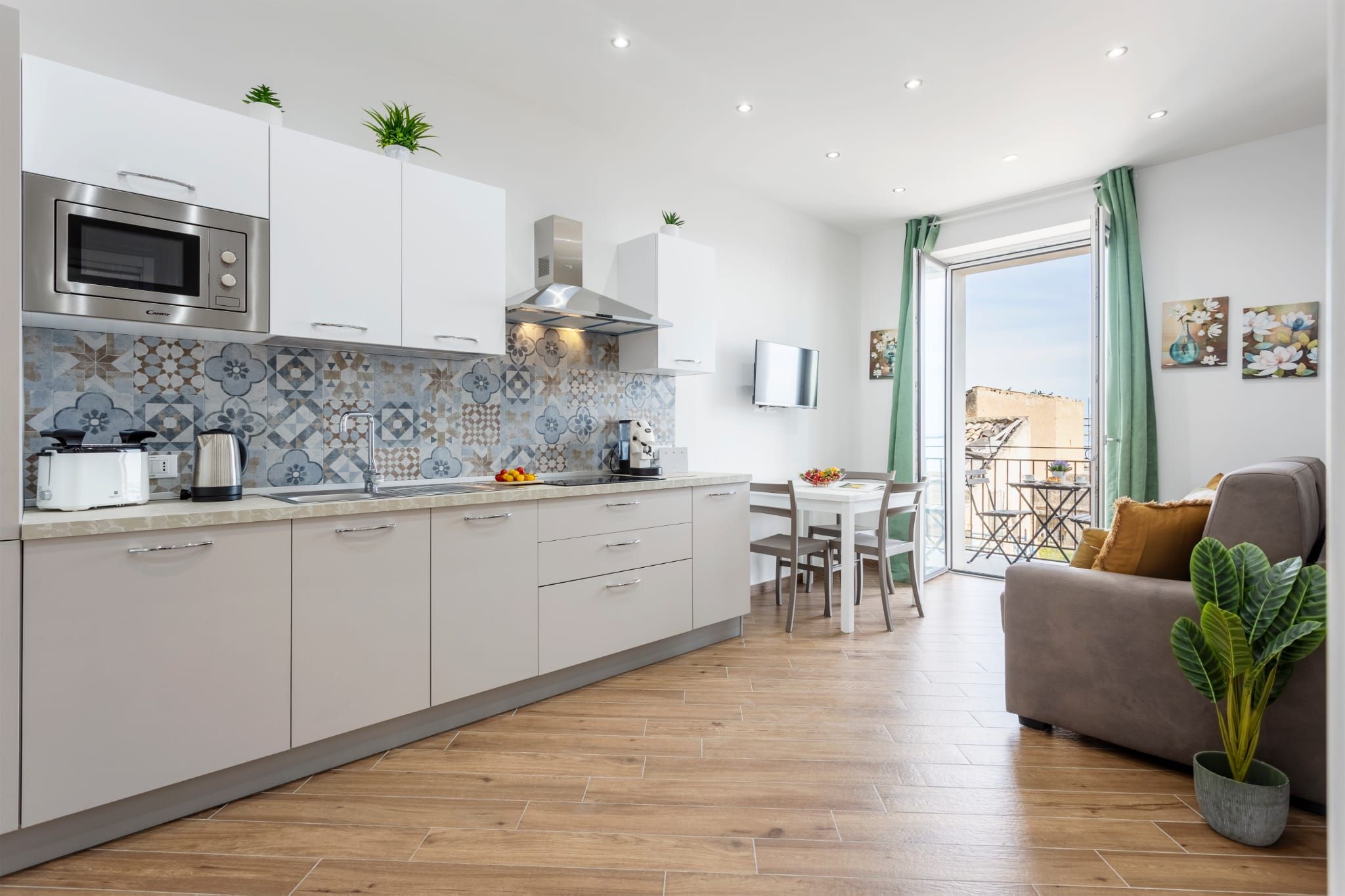 A modern kitchen with a microwave, toaster, and kettle, a dining area with a table and chairs, and a balcony with a view.