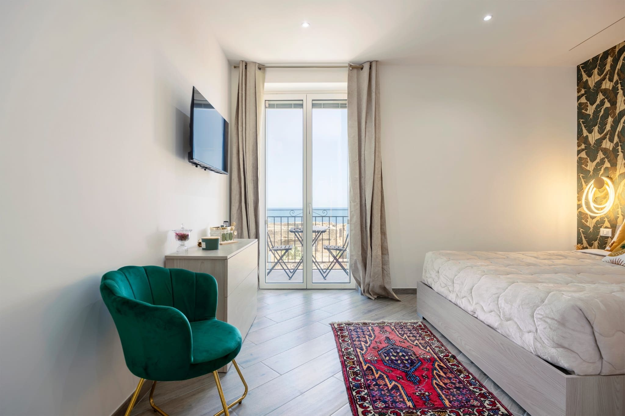 Habitación de hotel con cama, una silla verde, una mesita con objetos, un televisor montado en la pared y un balcón con mesa y sillas.