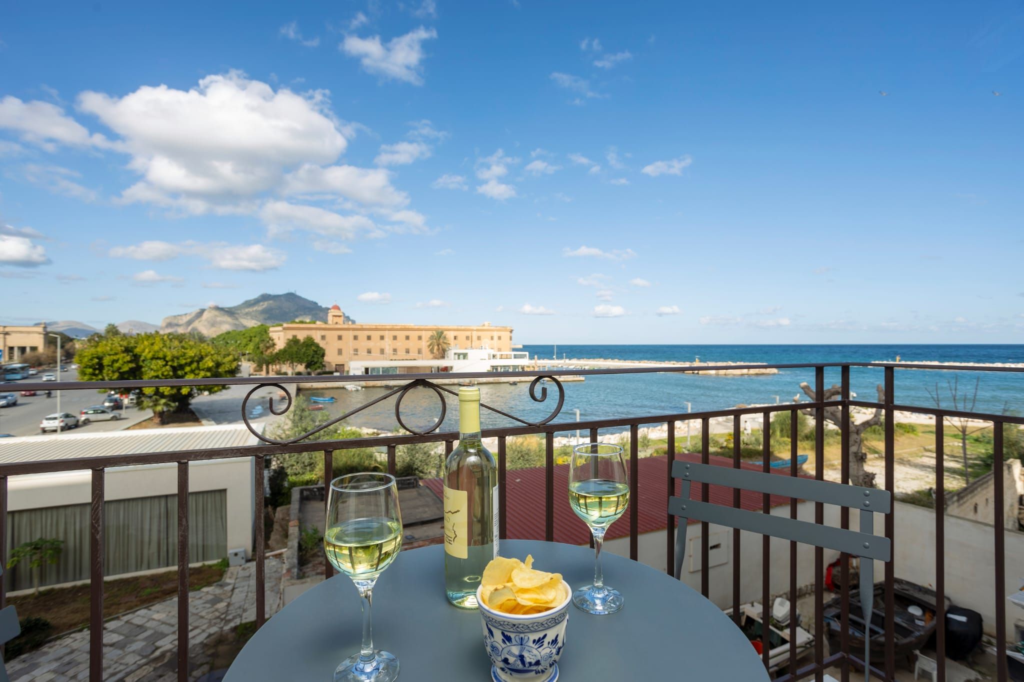 A balcony with a table set for two, overlooking a coastal view with a building in the background.