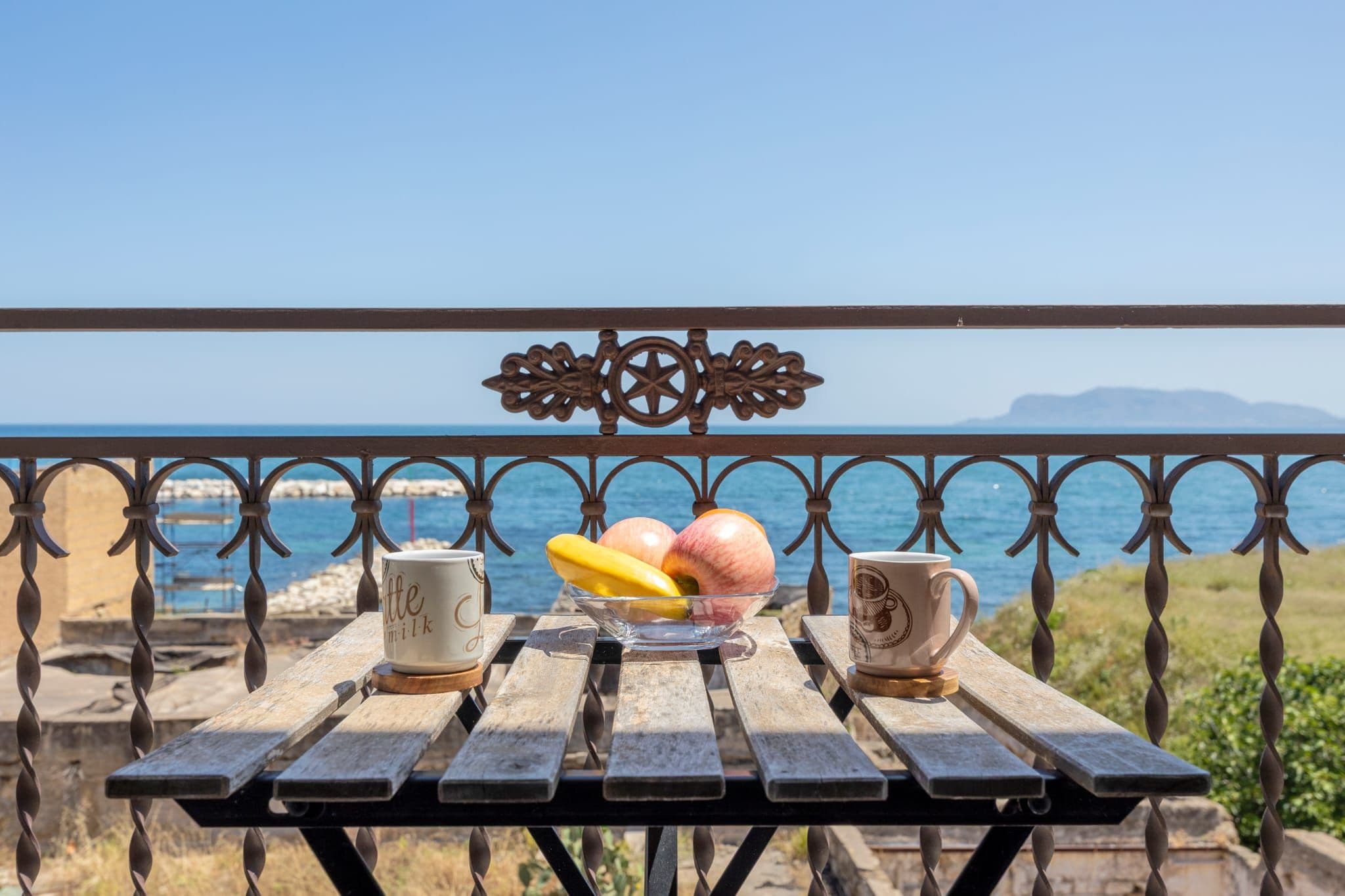 Une table en bois, avec deux tasses et un saladier de fruits, sur un balcon donnant sur la mer.