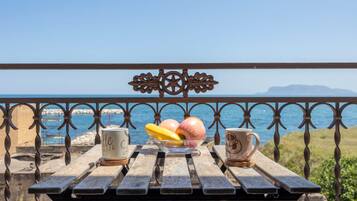 Une table en bois, avec deux tasses et un saladier de fruits, sur un balcon donnant sur la mer.
