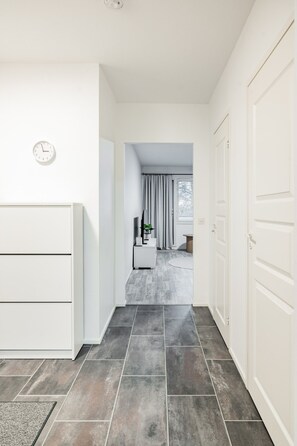 A hallway with a clock, a white cabinet, and a door leading to a room with a window and curtains. - Bright Nordic Apartment, Parking By Lapland Host (Rovaniemi)