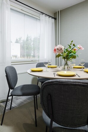 A dining area with a round table set for four, chairs, a vase with flowers, and a window with blinds. - Bright Nordic Apartment, Parking By Lapland Host (Rovaniemi)