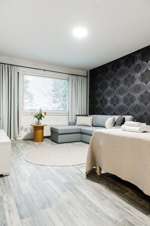 A modern bedroom with a sectional sofa, a bed, a round wooden side table, and a large window with curtains. - Bright Nordic Apartment, Parking By Lapland Host (Rovaniemi)