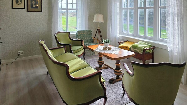 A room with a wooden table, four green upholstered chairs, a floor lamp, and a view of a green landscape through large windows.