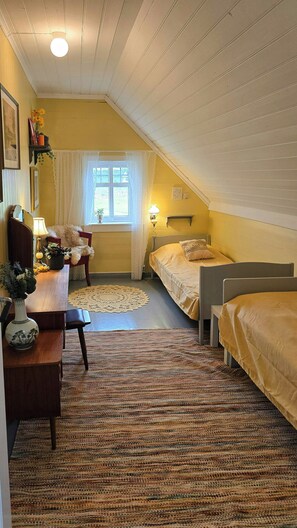 A cozy attic bedroom with a sloped ceiling, a desk with a lamp, two beds, a window with curtains, and a patterned rug.