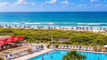 A pool area with lounge chairs and umbrellas, a beach with people and umbrellas, and a clear blue sky.