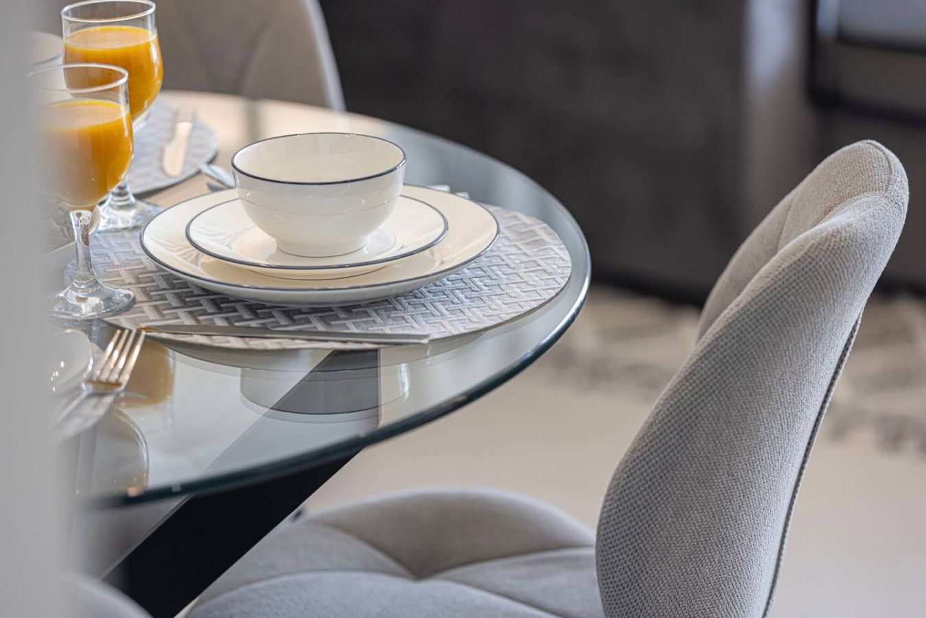 A dining table with a white bowl and saucer, a glass of orange juice, and a fork on a glass table.