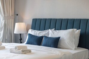 Een opgemaakt bed met witte lakens en twee blauwe kussens, een bedlampje en een gordijn.