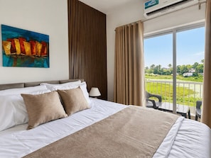 A hotel room with a large bed, a painting on the wall, a window with curtains, and a view of the outdoors. - Santorini Transit Apartment Negombo (Negombo)