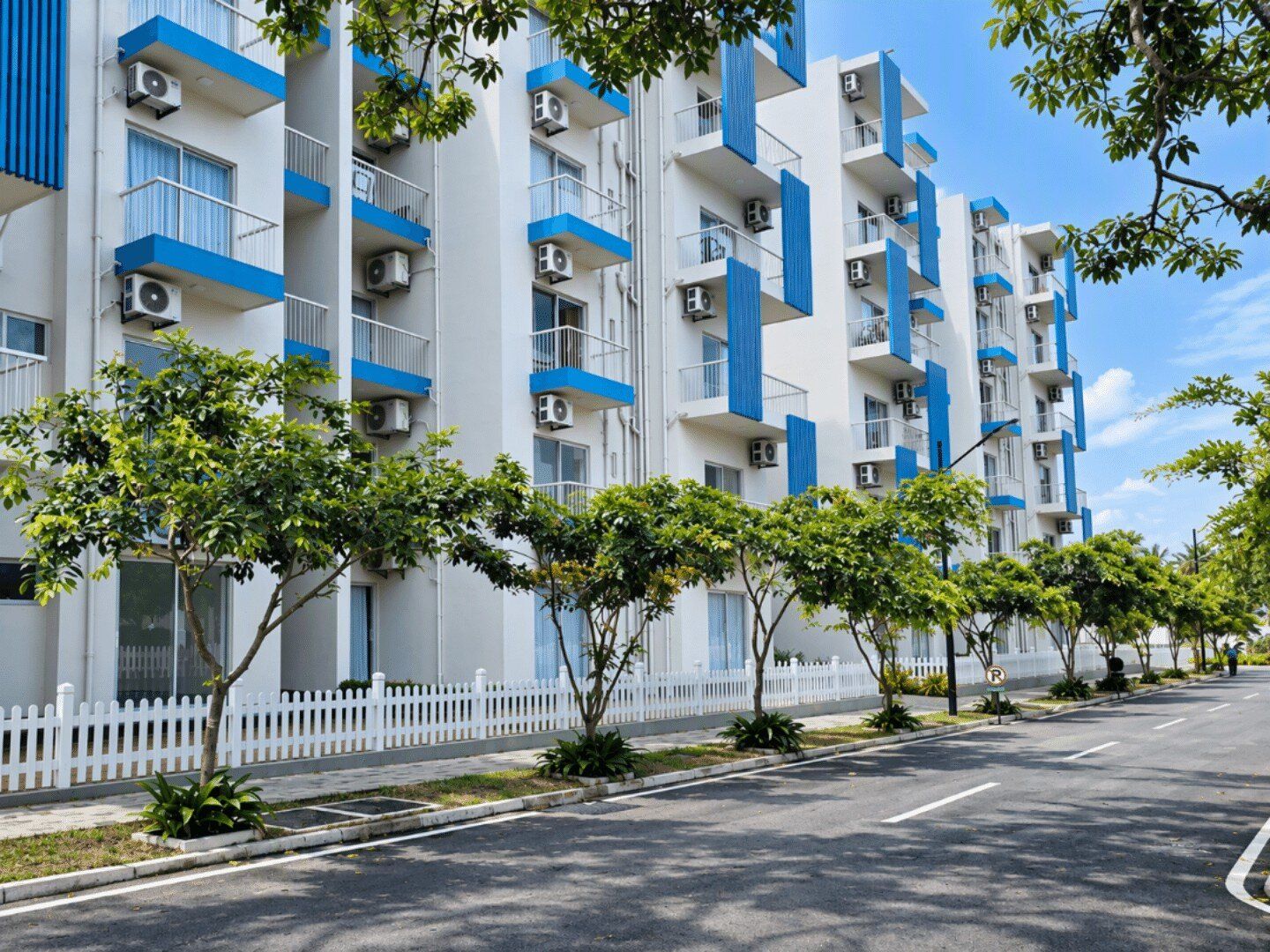 A multi-story residential building with balconies, air conditioning units, and a white picket fence.