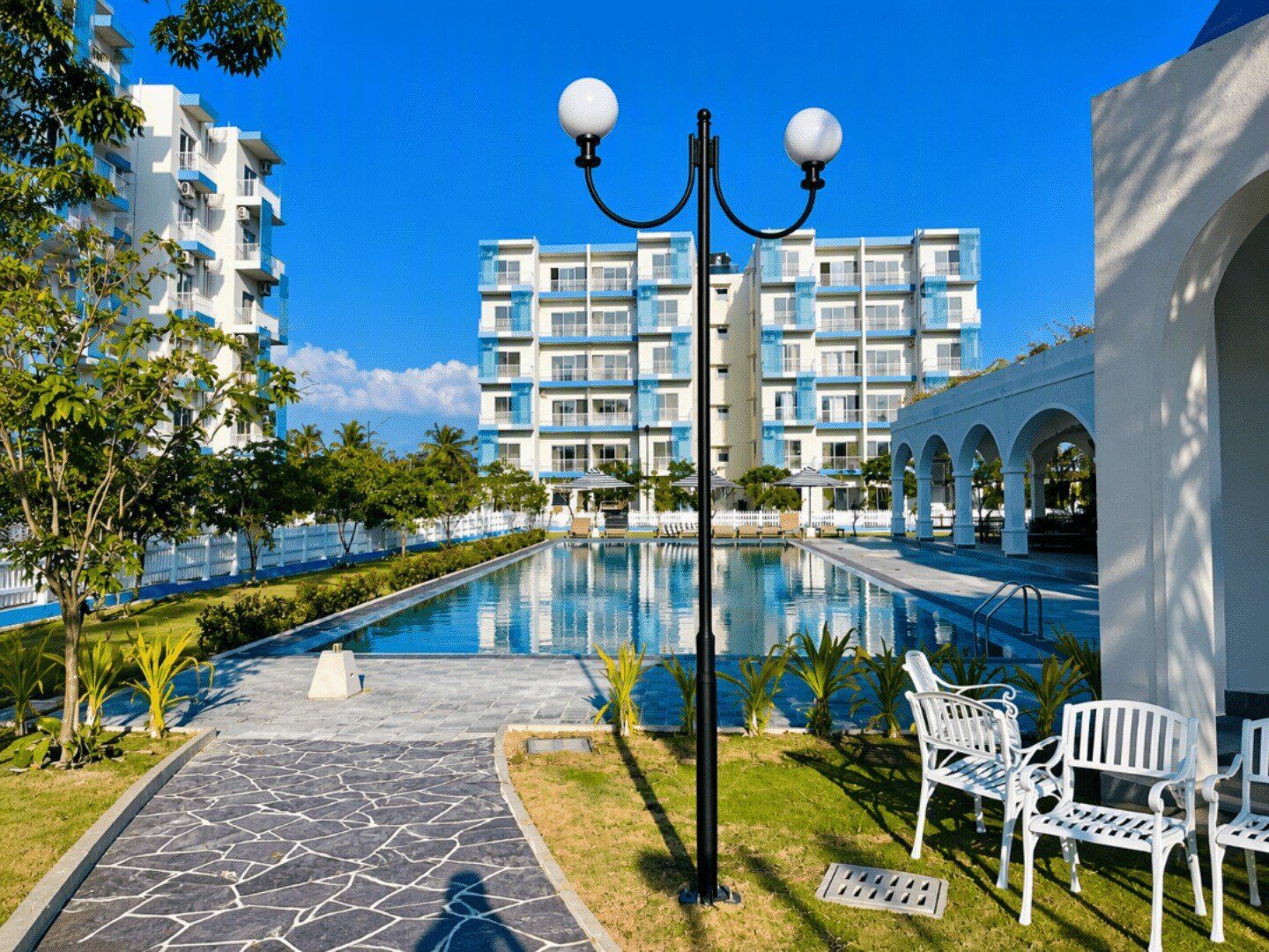 A swimming pool with a clear blue sky, surrounded by a white building with balconies, and a pathway leading to a seating area with white chairs.