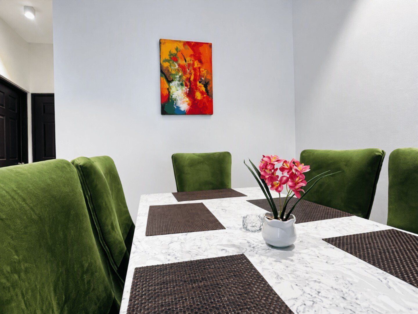A conference room with a white marble table, green upholstered chairs, a colorful abstract painting on the wall, and a vase with pink flowers.