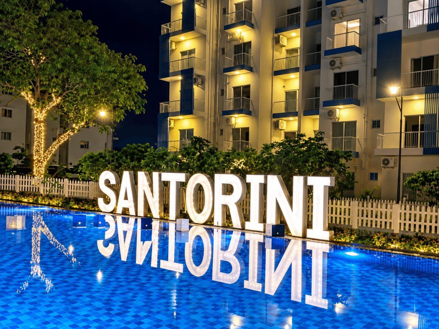 A swimming pool with the words 'SANTORINI INN' in large letters, surrounded by a white picket fence and trees with lights.