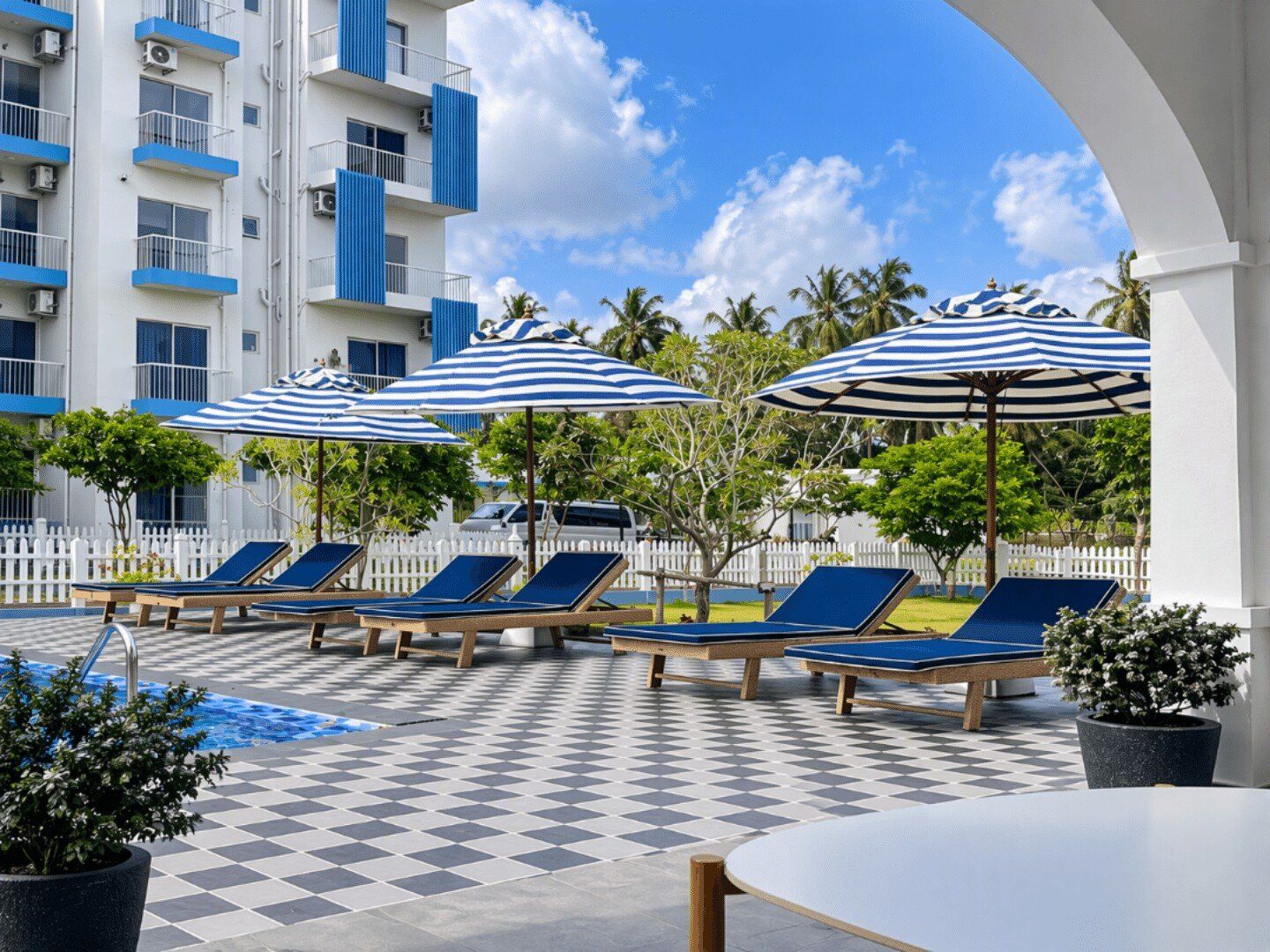 Un espace aménagé au bord de la piscine, avec des chaises longues et des parasols, un sol à carreaux et, au fond, un bâtiment doté de balcons.