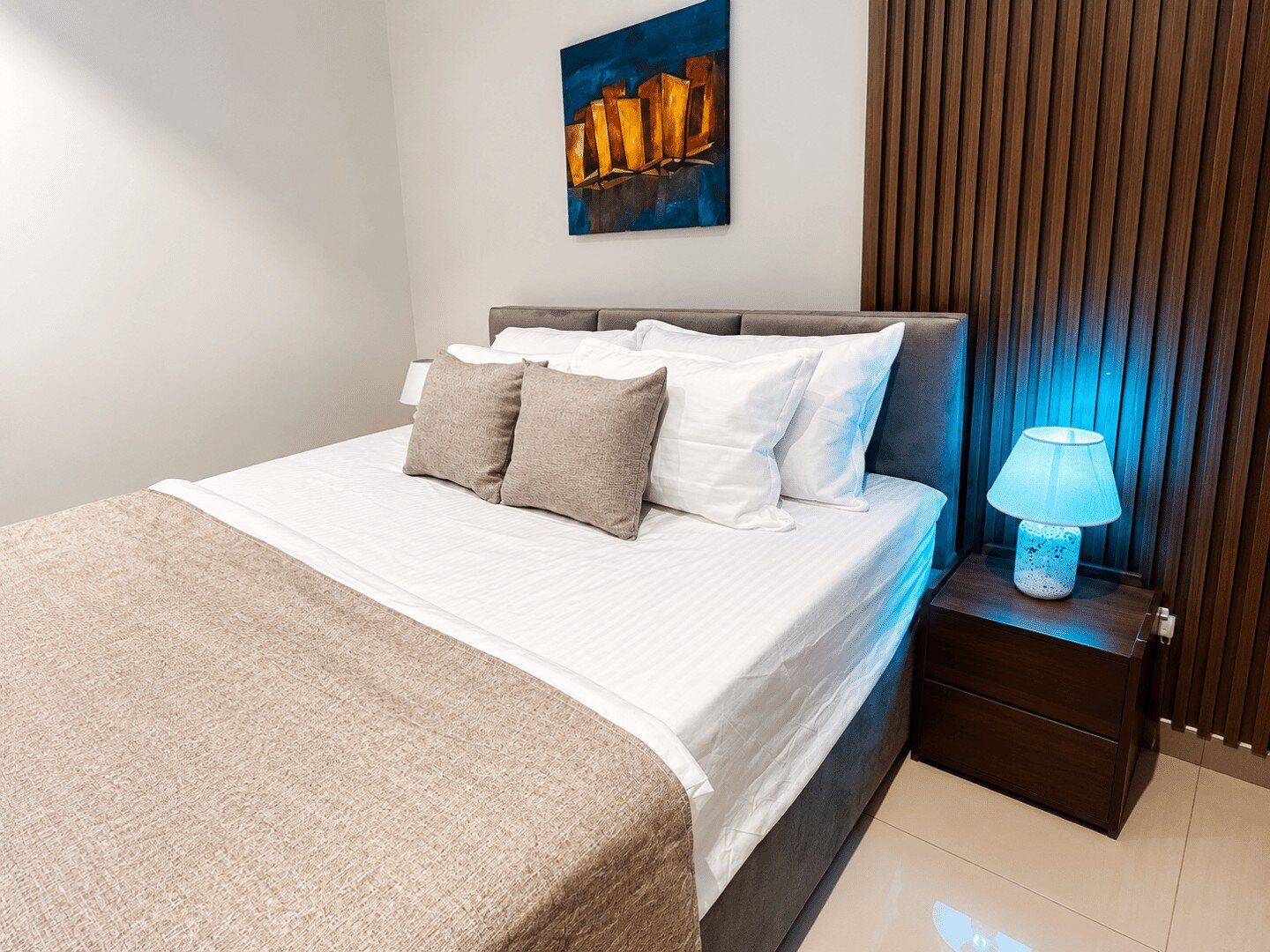 A neatly made bed with a white comforter and several pillows, a bedside lamp, and a wall-mounted painting.