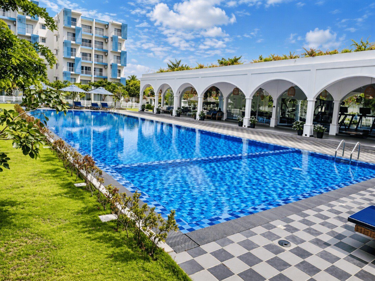 A swimming pool with a checkered pattern, surrounded by a covered area with arches, and a building in the background.