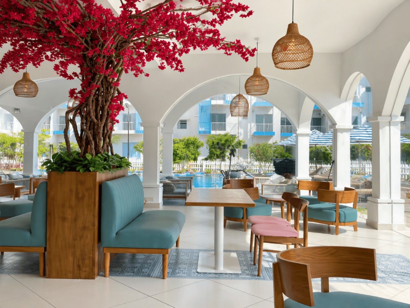 A spacious outdoor seating area with a large tree and hanging lights, overlooking a pool and buildings.