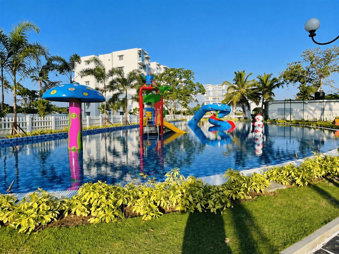 Una piscina con estructuras de juego coloridas, rodeada de palmeras y una cerca blanca.