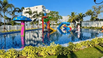 Una piscina con estructuras de juego coloridas, rodeada de palmeras y una cerca blanca.