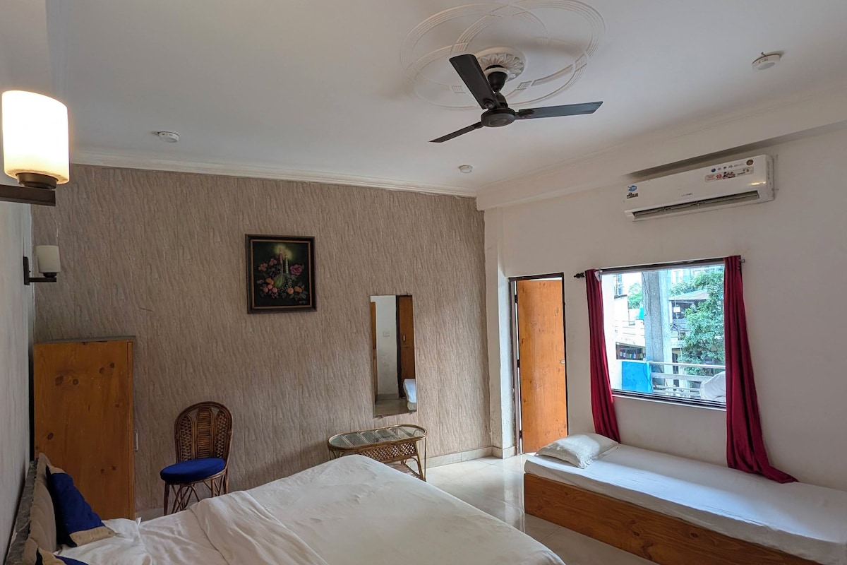 A bedroom with a bed, a chair, a small table, a window with curtains, and a ceiling fan.