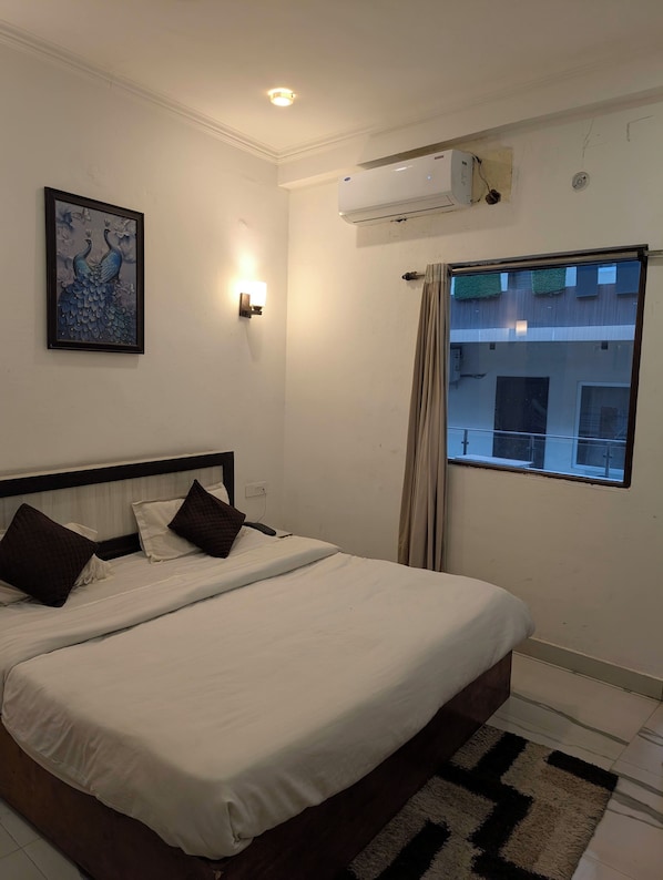 A hotel room with a bed, a wall-mounted air conditioner, a window with curtains, and a framed artwork on the wall.